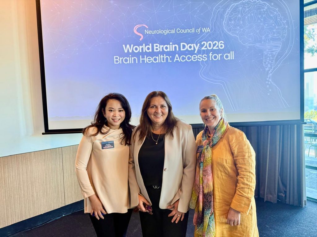 Alike WA team members Nadia and Nichola with a fellow attendee at the World Brain Day 2026 Breakfast Summit, standing in front of a presentation screen displaying the theme "Brain Health: Access for All".